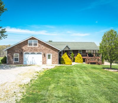 A brick and wood house with a green lawn, gravel driveway, garage, front porch, and trees under a clear blue sky in a rural setting.
