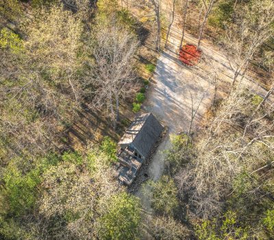 Aerial view of a small cabin surrounded by tall trees with early spring foliage. A dirt road runs nearby, and a red vehicle is parked beside the road among the trees. Sunlight filters through the branches.