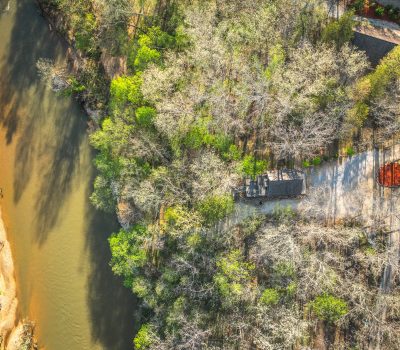 Aerial view of a winding river beside a forest with both green and leafless trees; a small building and a driveway are visible near the water’s edge.