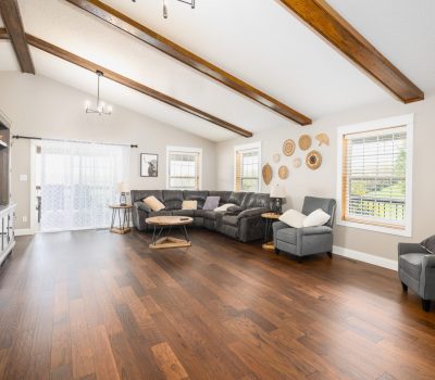 A spacious living room with wooden floors, exposed ceiling beams, a large sectional sofa, armchairs, a TV unit, round coffee tables, wall art, and large windows letting in natural light.
