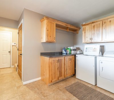 A laundry room with tile floor, wooden cabinets, a black countertop, a washing machine, a dryer, and laundry supplies; a hallway with a white door is visible to the left.