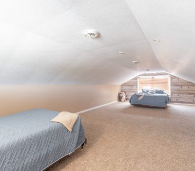 A narrow attic bedroom with sloped ceilings, two single beds with blue bedding, beige carpet, and a window at the far end letting in natural light.