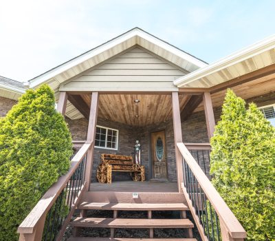 A front porch with wooden steps, a brown bench, and a decorative glass door, flanked by two trimmed evergreen bushes and a house exterior of stone and siding under a peaked roof.