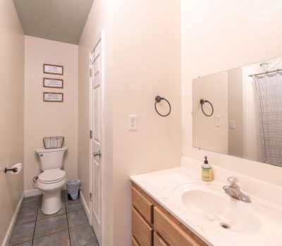 A bright, narrow bathroom with a sink, mirror, and wooden cabinet on the right. At the end, there is a toilet with wall art above it, a waste bin, and a shower with a white curtain. Walls and tiles are light-colored.
