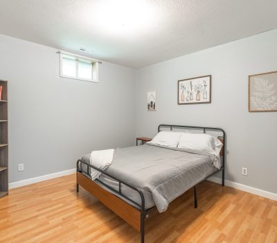 A neatly made bed with gray bedding is centered in a simple bedroom with light gray walls, wood flooring, a bookshelf, and framed art on the walls. A small window lets in natural light above the bed.