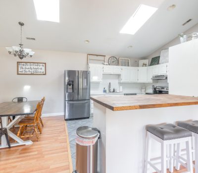 Bright kitchen and dining area with white cabinets, stainless steel appliances, wooden dining table with mix of chairs, bar counter with two stools, and wall decor. Light wood flooring and skylights add natural light.