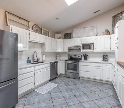 A spacious kitchen with white cabinets, stainless steel appliances, a tiled floor, a large refrigerator, and a skylight. Decorative items and plants are placed on top of the cabinets.