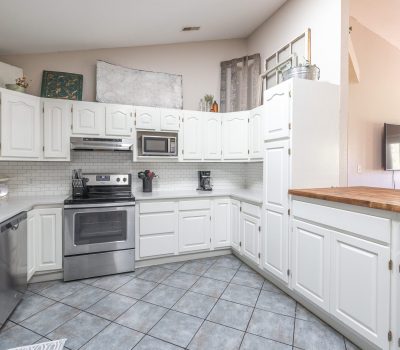 A bright, spacious kitchen with white cabinets, stainless steel appliances, subway tile backsplash, tiled floor, and a large wooden countertop on the right. Decor and plants are placed above the upper cabinets.