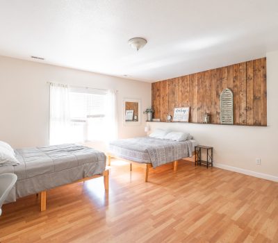 A bright bedroom with two beds, wood floors, a wooden accent wall behind one bed, a window with sheer curtains, and minimal decor including a mirror, framed art, and plants.