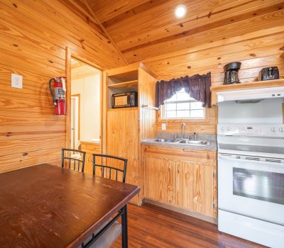 A small kitchen with wood-paneled walls and ceiling, featuring a white stove, oven, microwave, sink, coffee maker, and a dark wooden dining table with chairs. Natural light enters through a window above the sink.