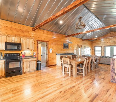 A spacious, open-concept cabin kitchen and dining area with wooden walls, floors, and cabinets. Black appliances, a large wooden dining table, and a living area with a fireplace and sofas can be seen. The ceiling is metal with exposed beams.