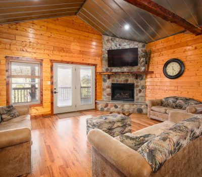 A cozy log cabin living room with wooden walls, three patterned sofas, a stone fireplace with a TV above it, a clock on the wall, large windows, and glass double doors opening to a deck.