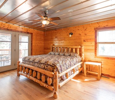 A cozy wooden bedroom with a rustic log bed, patterned bedding, wooden walls, hardwood floors, a ceiling fan, large windows, and glass doors leading to a balcony. Sunlight brightens the room.
