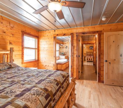 A cozy log cabin bedroom with wood-paneled walls and ceiling, a rustic wooden bed with a patchwork quilt, a ceiling fan, and doorways leading to a bathroom and other rooms. Natural light streams through a window.