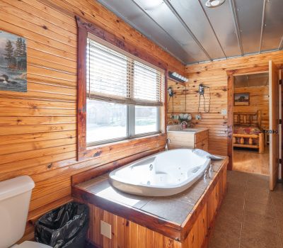 A rustic bathroom with wood-paneled walls, a large soaking tub, a toilet, a window with blinds, a vanity with a sink, and an open door leading to a bedroom with a wooden bed.