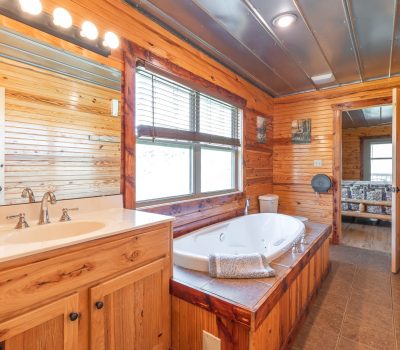 A rustic bathroom with wood-paneled walls, a large corner bathtub with towels, a sink with a mirror, and a window letting in natural light. An open door reveals a glimpse of a sitting area beyond.