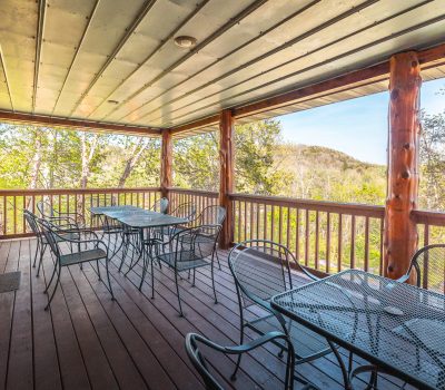 Covered wooden deck with three metal patio tables and chairs, surrounded by a wooden railing. The deck overlooks green trees and distant hills under a clear sky, creating a peaceful outdoor setting.