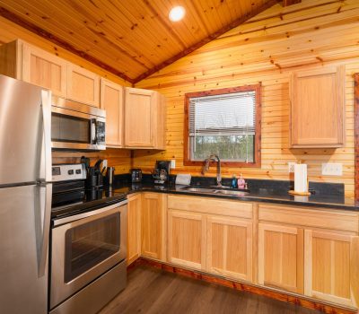 A cozy kitchen with wooden cabinets and walls, stainless steel appliances, a sink under a window, and various small kitchen appliances on the black countertop. Natural light comes in through the window.