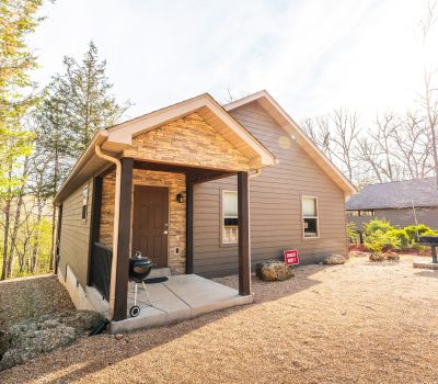 A small brown cabin with a covered front porch, surrounded by trees and gravel. Sunlight filters through the foliage, and a "For Sale" sign is visible near the front of the house.