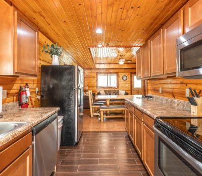 A cozy log cabin kitchen with wood-paneled walls and ceiling, brown cabinets, stainless steel appliances, and a dining area with a wooden table and benches in the background.