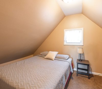 Small attic bedroom with beige walls and sloped ceiling, featuring a bed with gray bedding, a single pillow, a window above the bed, and a black nightstand with a lamp. Brown carpet covers the floor.