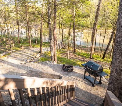 View from a wooden staircase overlooking a gravel patio area with a fire pit and a black barbecue grill, surrounded by trees, near a river in a forest setting on a sunny day.