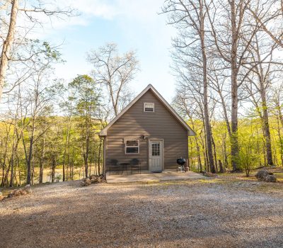 A small brown cabin with a gabled roof sits among tall, leafless trees. There are two black chairs, a small table, and a grill in front of the porch, with gravel and sparse green foliage surrounding the cabin.