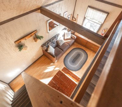 View from a staircase looking down into a cozy living room with a brown couch, white pillows, a woven rug, wooden furniture, a window with a curtain, and wall-mounted decorations. Sunlight streams in from the window.