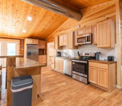A spacious kitchen with wood-paneled walls and ceiling, stainless steel appliances, light wood cabinets, a center island with stools, and double glass doors letting in natural light.