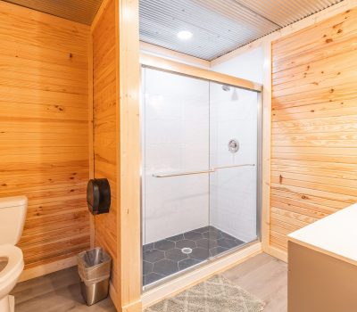 A modern bathroom with light wooden walls, a white toilet, a glass-enclosed shower with dark hexagon tiles, a small white sink, gray bath mat, metal trash can, and bright overhead lighting.