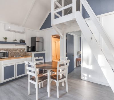 A small, bright kitchen and dining area with white cabinets, a wooden table with four chairs, a loft staircase, a microwave, a fridge, and a coffee maker under high, white ceilings.
