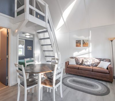 Bright, modern living space with a brown sofa, round dining table with four chairs, a loft area accessed by stairs, wall art, tall lamp, and a window allowing natural light into the room.