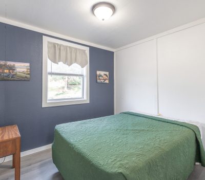 A small, bright bedroom with white walls, one blue accent wall, a window with a valance, a double bed with a green quilt, a wooden nightstand with a lamp, and two landscape paintings on the wall.