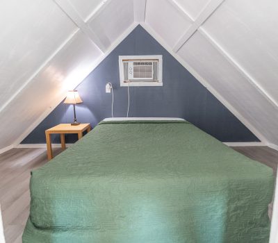 A cozy attic bedroom with a slanted white ceiling, blue accent wall, a bed with a green quilt, a small wooden nightstand with a lamp, and a window air conditioner above the bed.