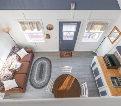 A top-down view of a cozy, bright living space with a brown sofa, round dining table with four chairs, small kitchen area, and a blue door. Light streams in through two windows, illuminating the tidy, modern room.