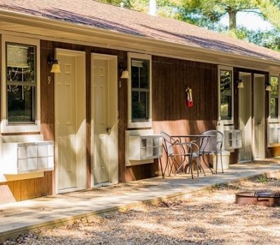 A row of attached cabins with brown wooden siding and tan doors. Each unit has a small porch area with two metal chairs and a table. The front yard has a gravel area and a small fire pit. Trees in the background suggest a woodland setting.