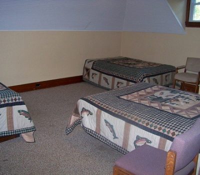 A bedroom with three single beds covered in bedspreads featuring fish patterns. The room has beige walls, a slanted ceiling, two wooden chairs, and a small window near the ceiling. The floor is carpeted, and the furniture is simple and wooden.